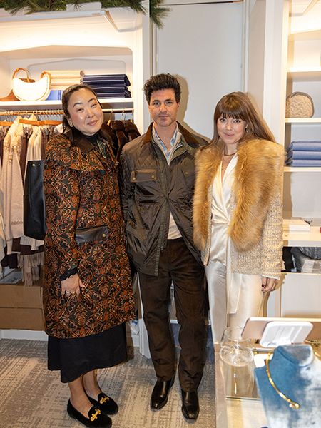 Three people in formal attire pose together at an upscale holiday event or party. Holiday decorations including garland are visible in the background of what looks like a retail or boutique setting.