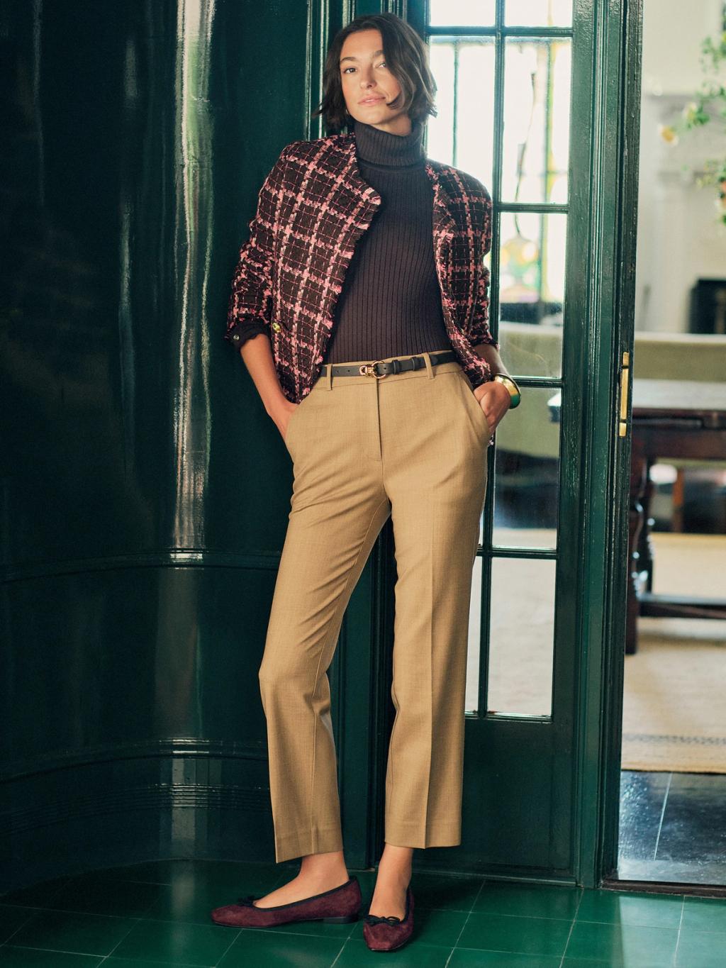 A woman in a brown ribbed turtleneck, pink and brown plaid tweed jacket, khaki pants, and burgundy ballet flats leans against a dark green door.