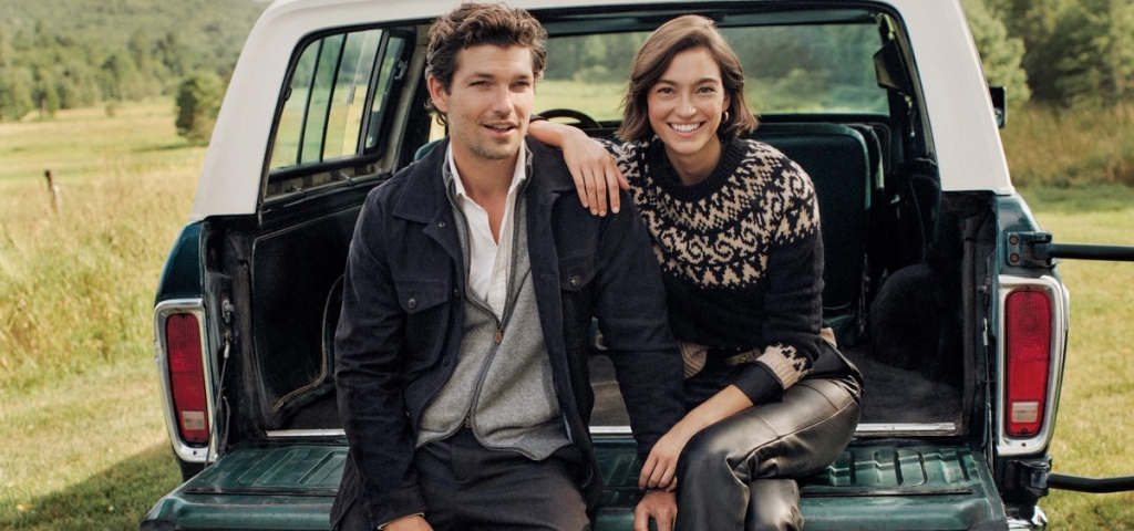A man and woman sit smiling on the tailgate of a truck in a rural setting.