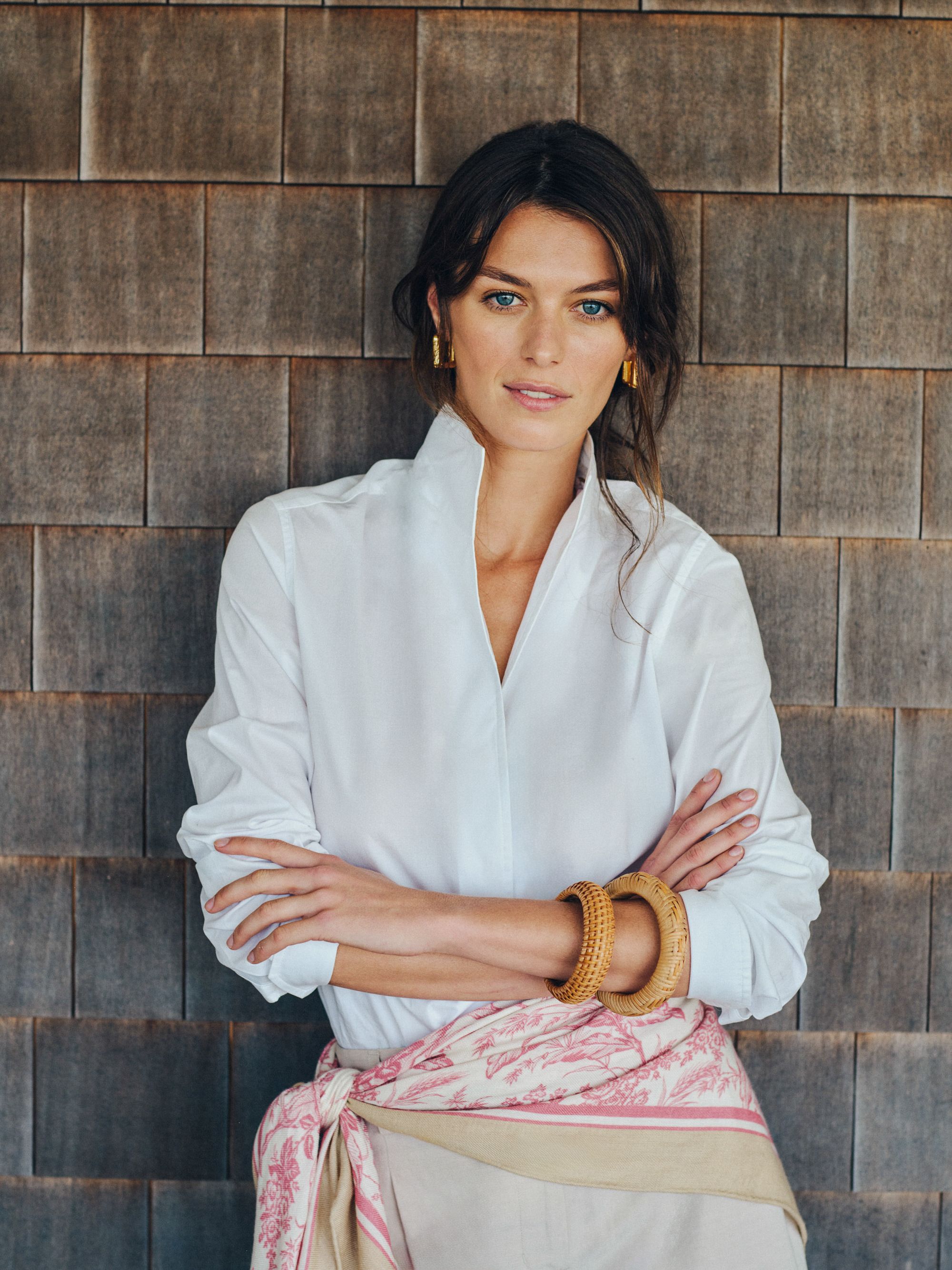 A woman in a white collared shirt, pink patterned scarf, and wooden bangles stands with crossed arms against a shingled wall.