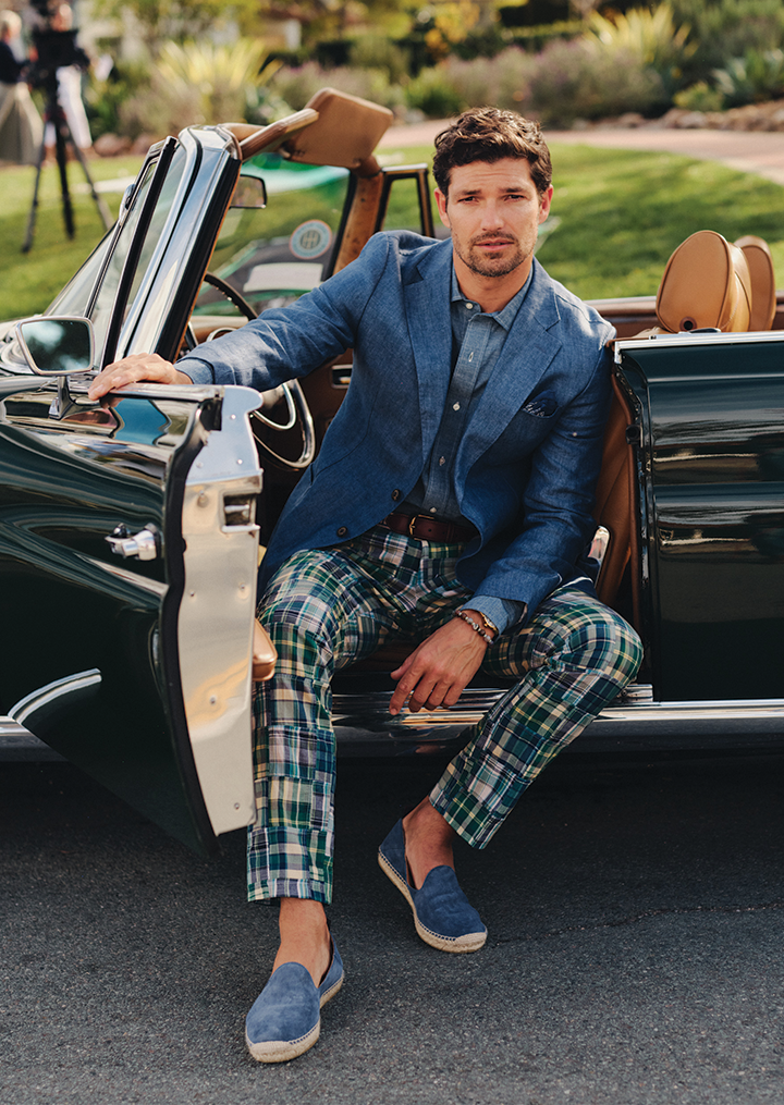 A man in a blue linen jacket, patchwork plaid pants, and blue espadrilles sits on the edge of a vintage green convertible, looking at the camera.