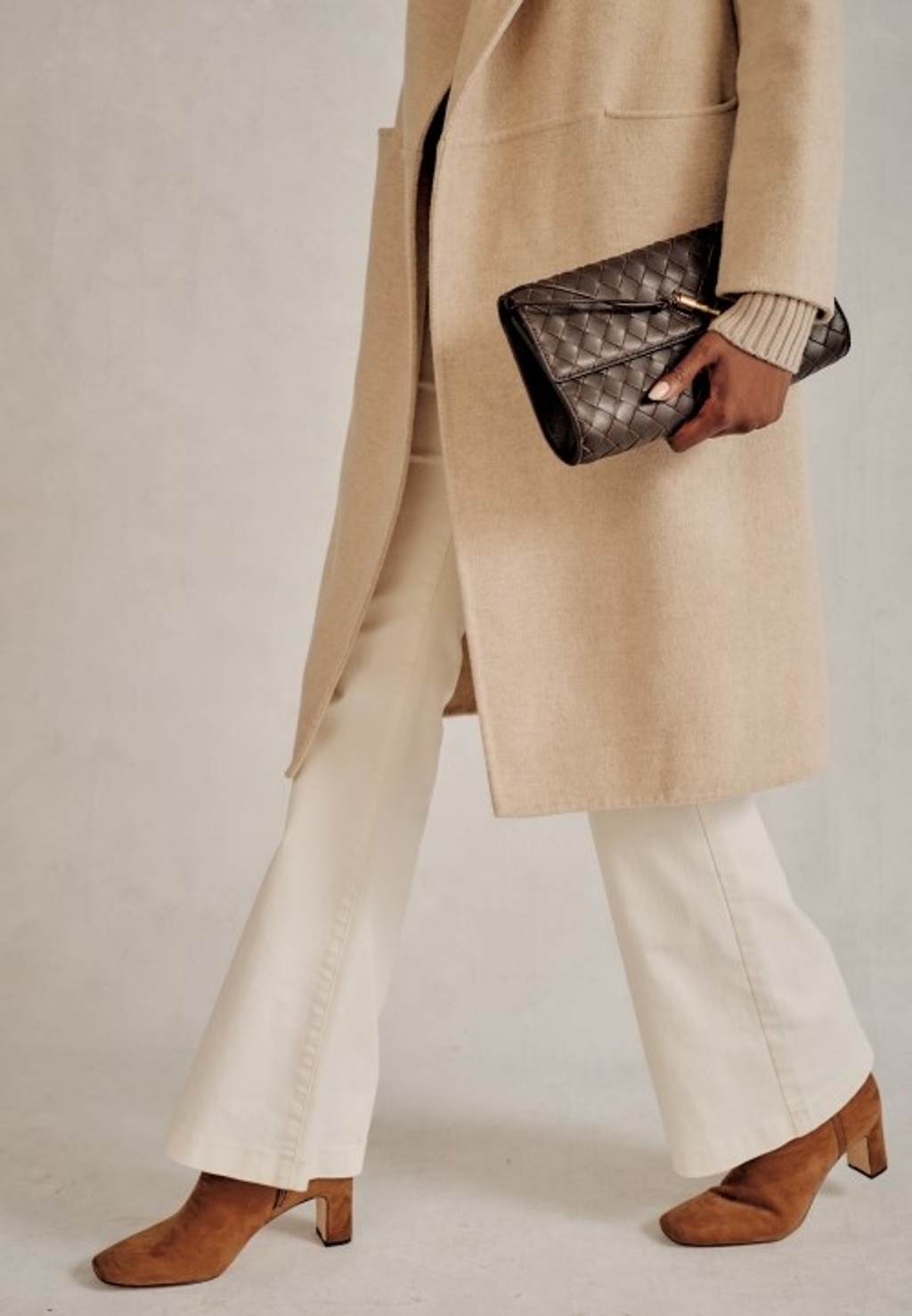 Women with brown heeled boots, white pants, and a cream colored trench coat walking while holding a brown leather clutch.