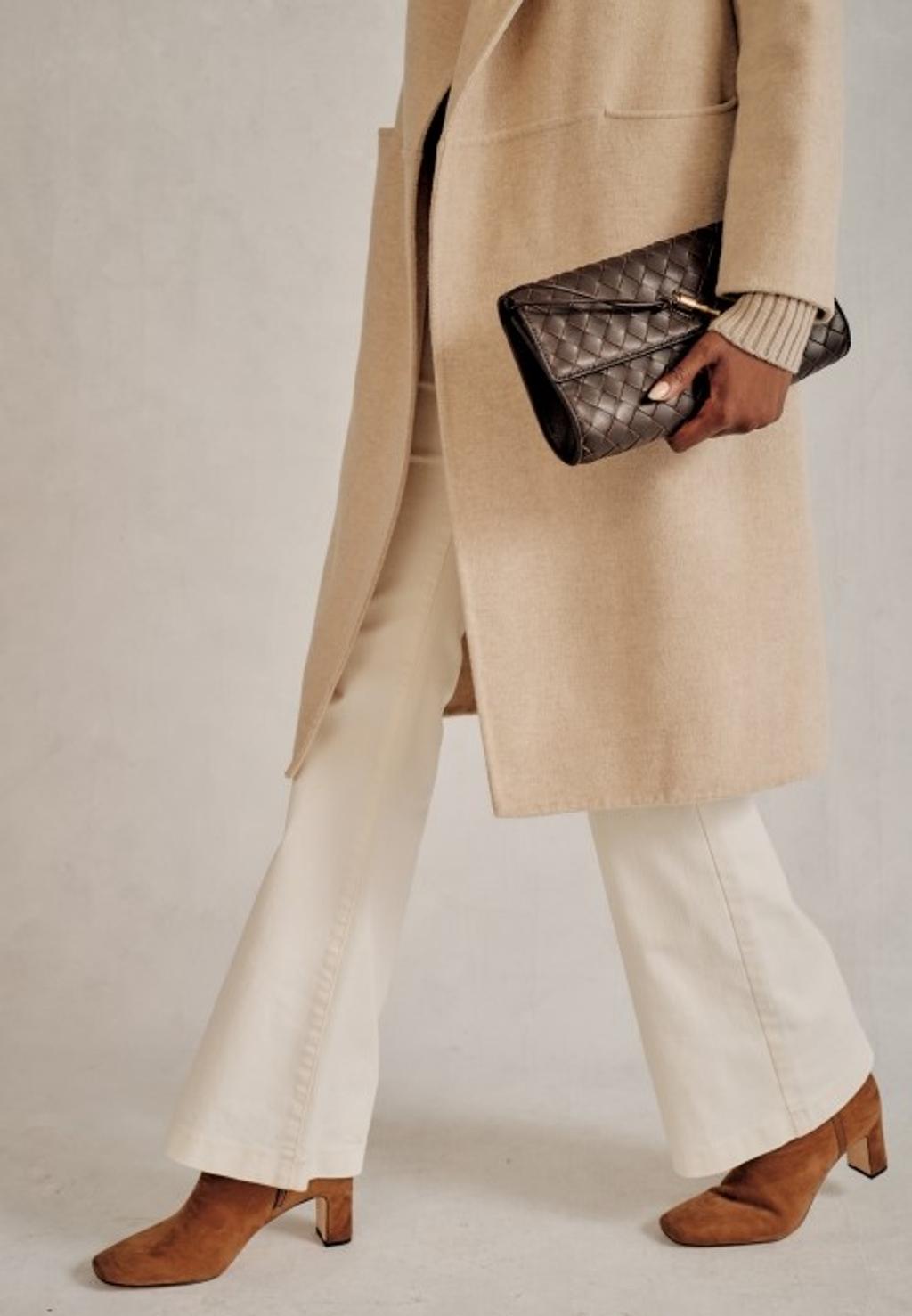 Women with brown heeled boots, white pants, and a cream colored trench coat walking while holding a brown leather clutch.
