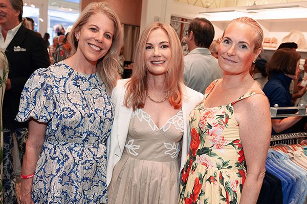 Three smiling women in a blue patterned dress, a beige dress with a white blazer, and a yellow floral dress pose indoors.