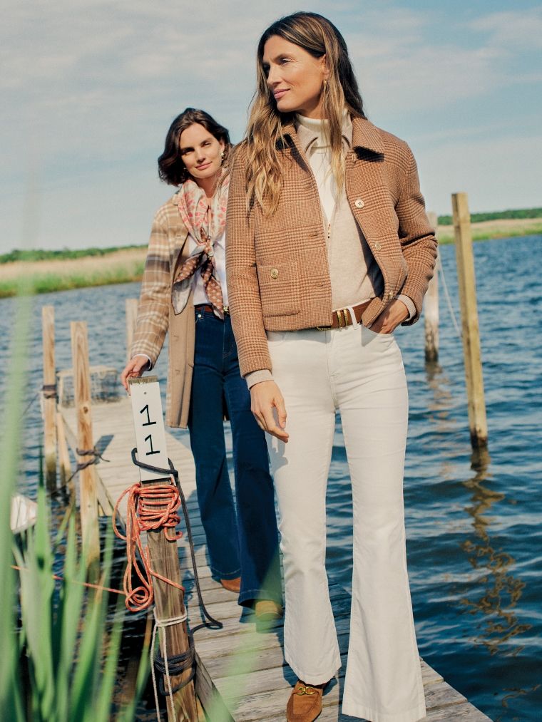 Two women walk on a wooden dock by the water; one in a brown plaid jacket and white flared pants, the other in a plaid coat and patterned scarf.