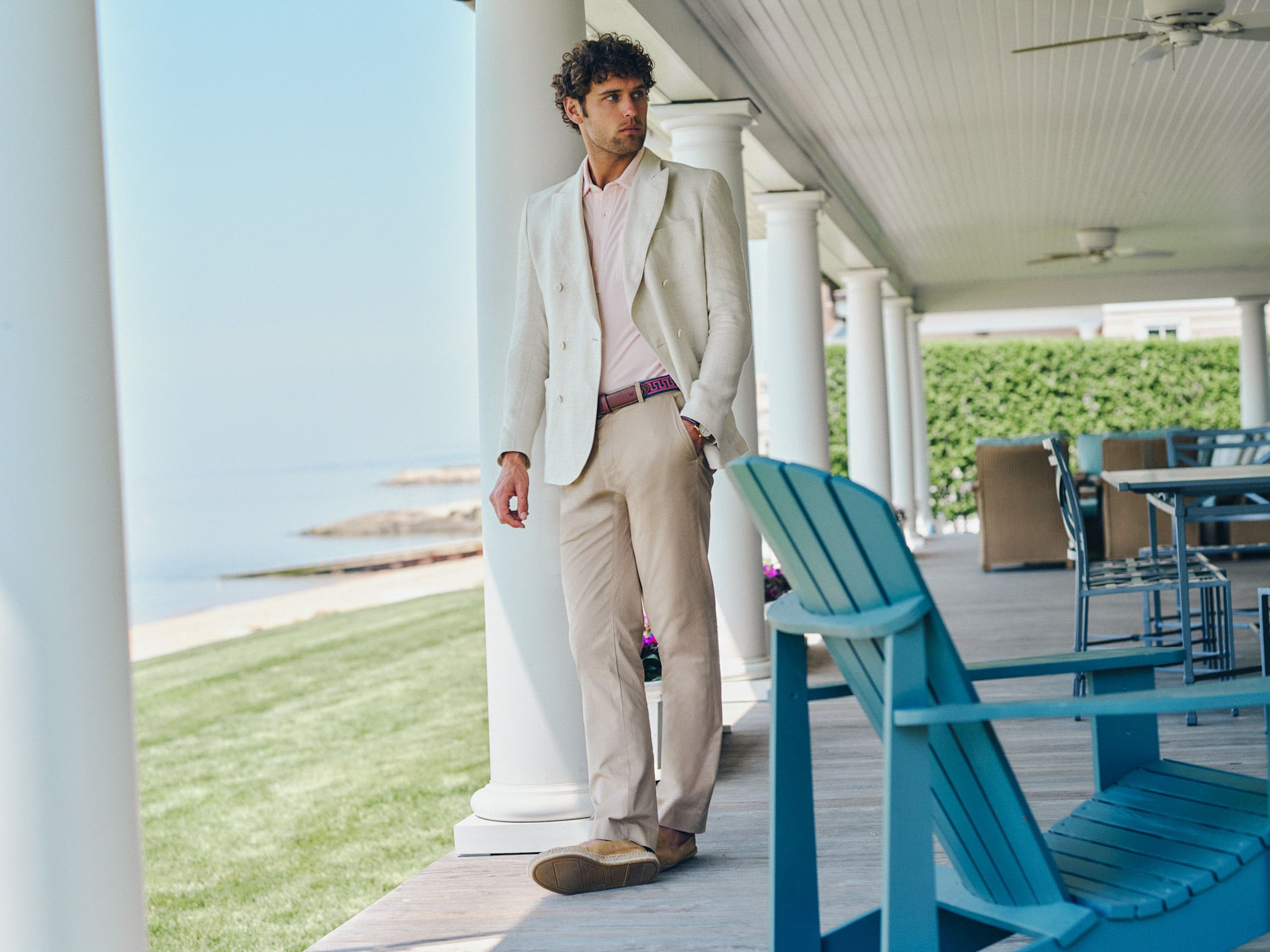 A man in a cream suit and pink shirt stands on a white-pillared porch overlooking the ocean.