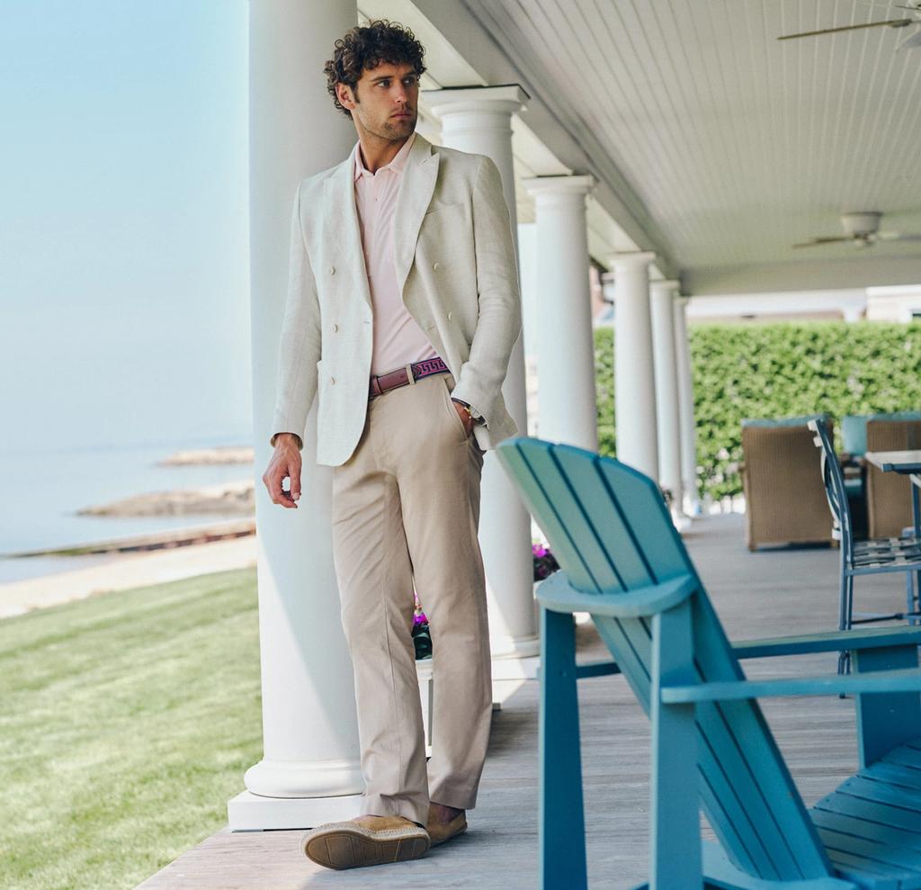 A man in a cream suit and pink shirt stands on a white-pillared porch overlooking the ocean.