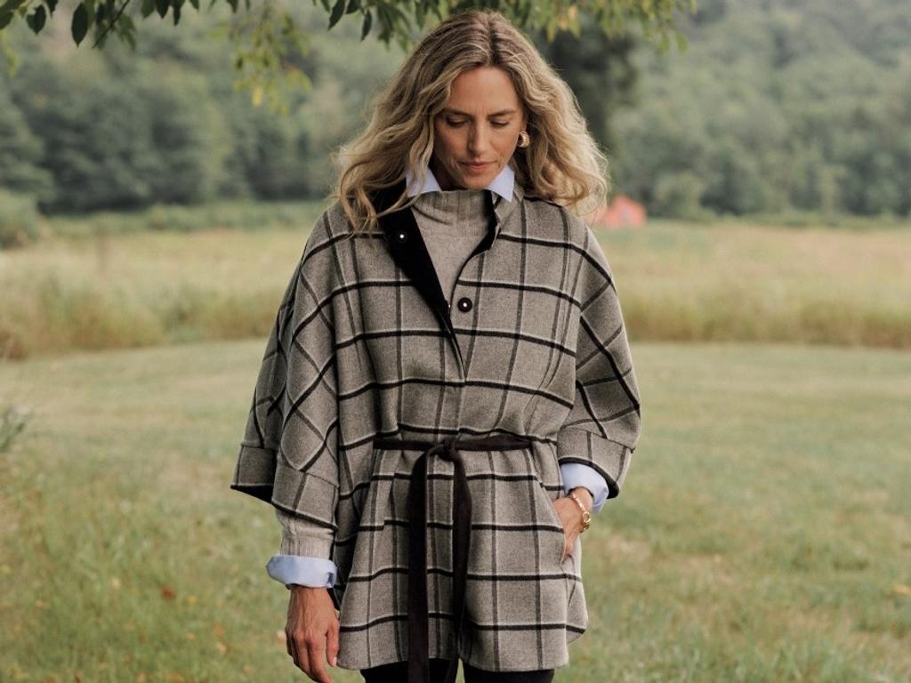A woman wearing a grey plaid belted poncho over a light blue shirt, looking down outdoors.