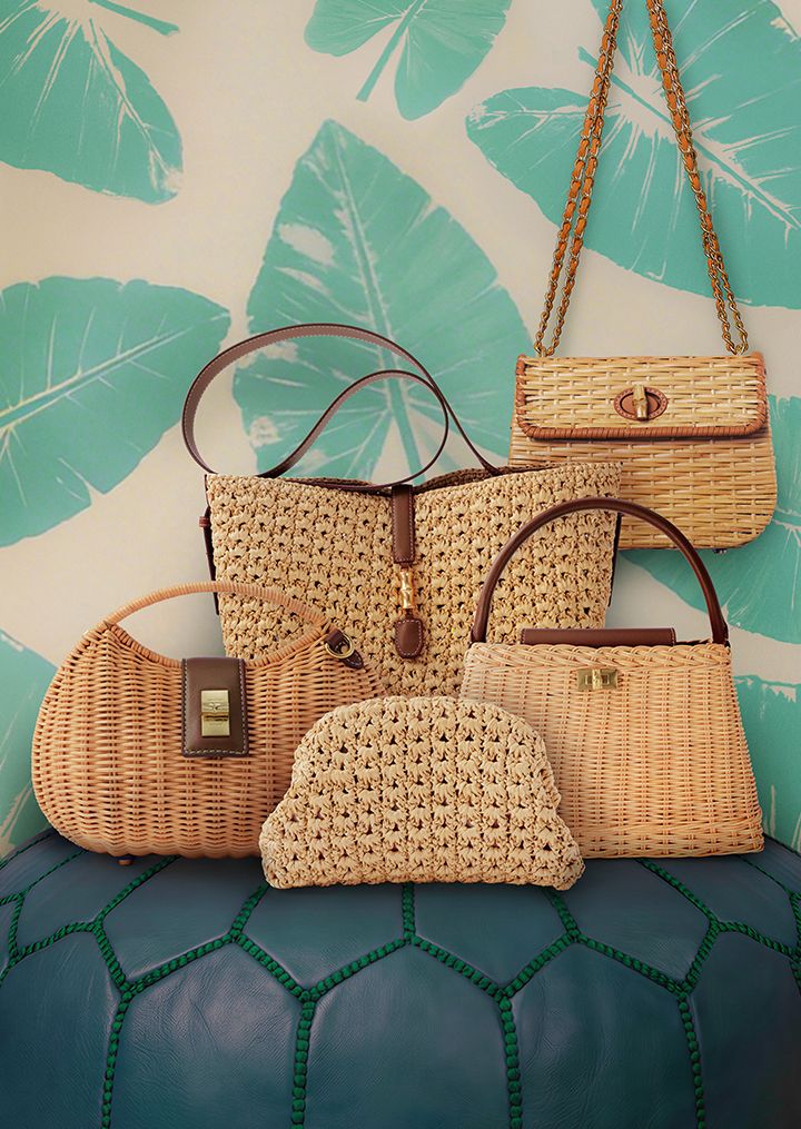 A collection of five woven straw and wicker bags with brown leather accents, displayed against a wall with turquoise leaf patterns, sitting on a dark blue textured surface.