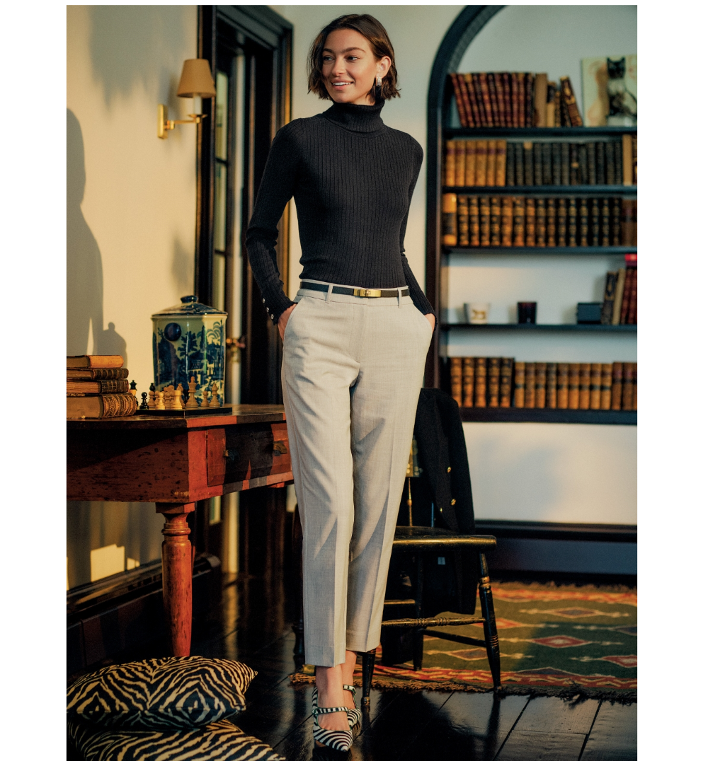 A smiling woman in a black ribbed turtleneck, grey trousers, and zebra-print heels stands in a library-like room.