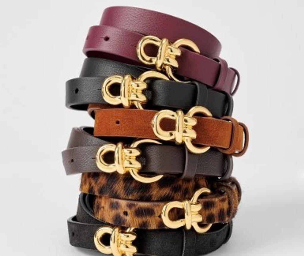 A stack of belts in various colors and materials, including burgundy, black, brown, and leopard print, each with a gold double-loop buckle.