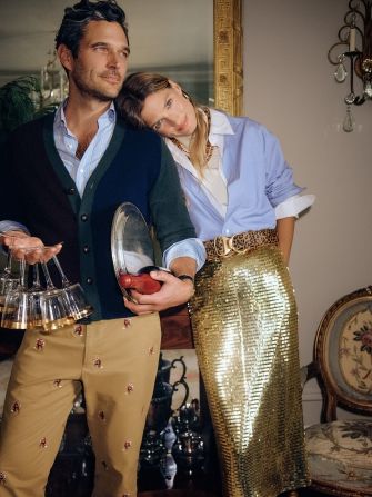 man and woman standing in living room party scene, she is wearing blue button down shirt and gold sequin skirt