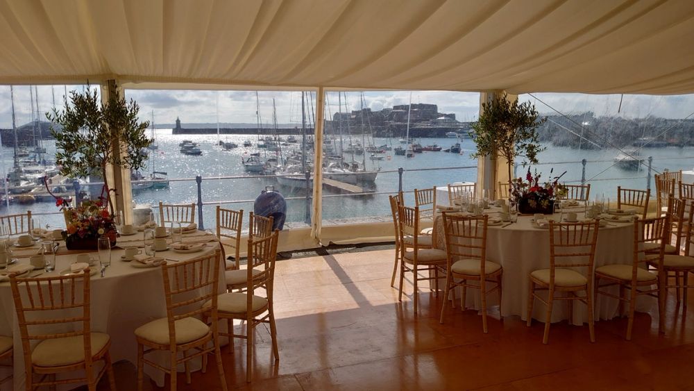 Gold chairs and dressed tables under a marquee with the harbour and Castle Cornet beyond