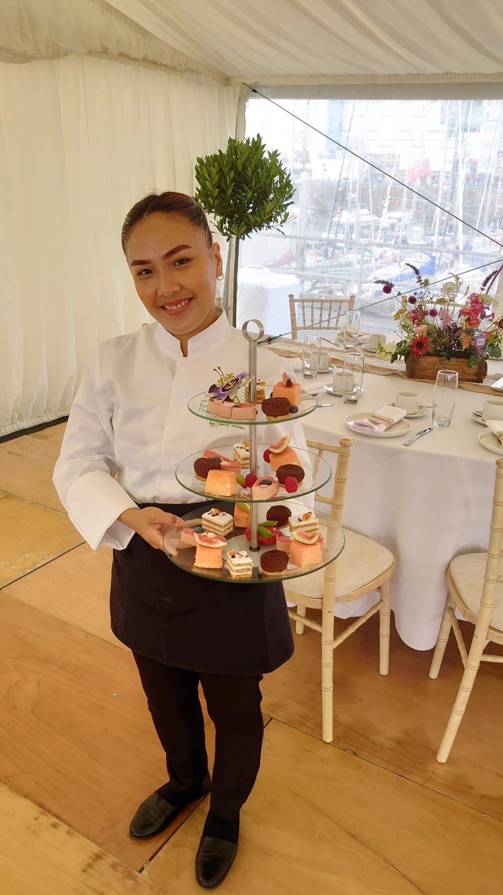 A Catered by Randalls server in chef whites presenting a tiered afternoon tea inside a harbour-side marquee