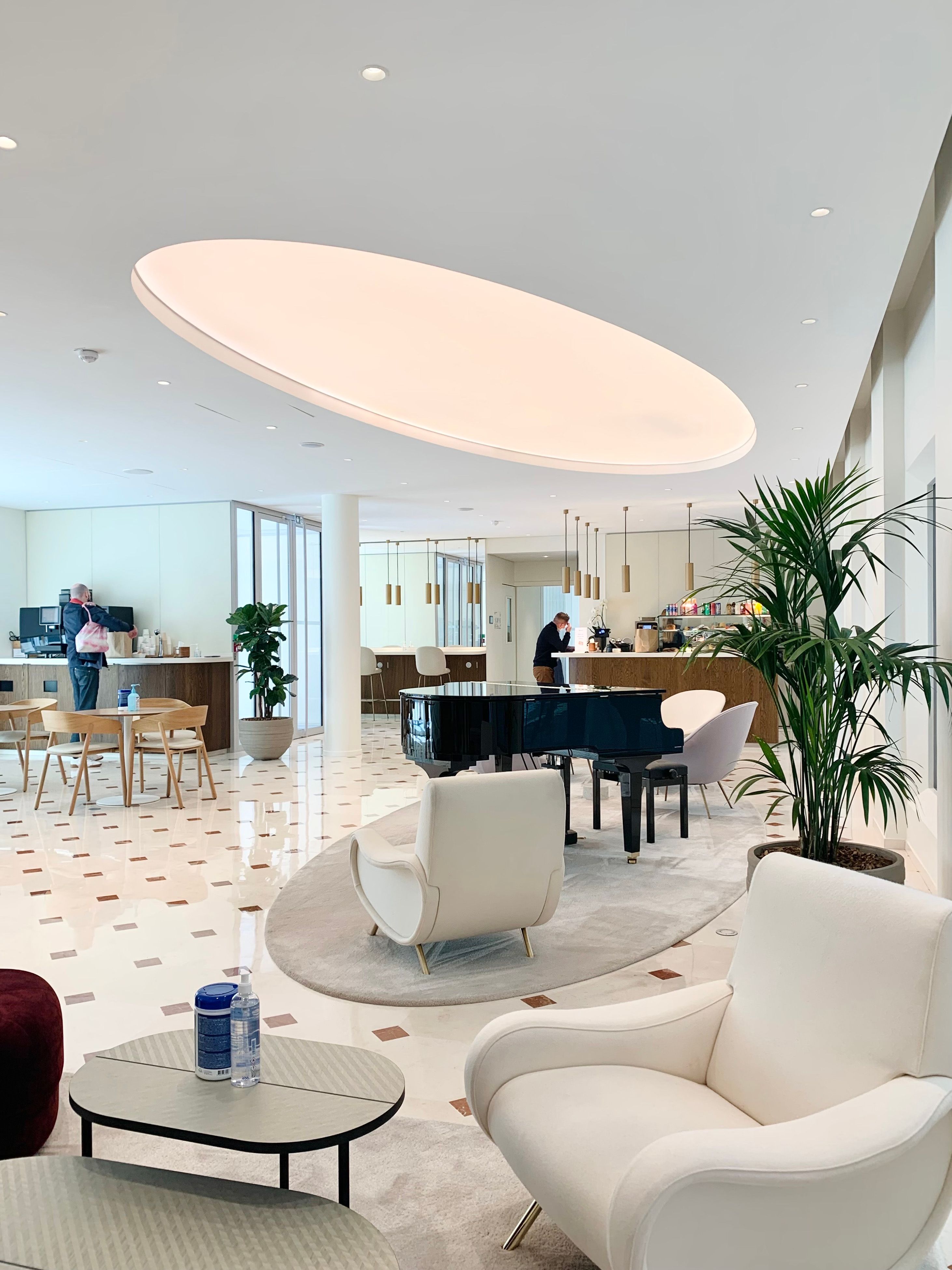 Moderne Innenarchitektur mit einer zentralen Lichtdecke, unter der sich ein schwarzer Flügel befindet, umgeben von eleganten Sesseln