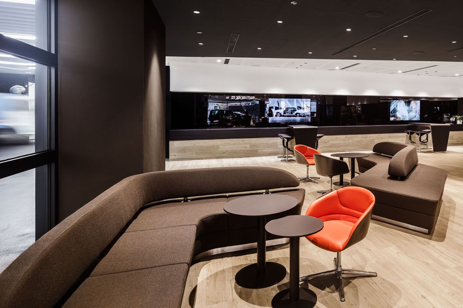 Moderne Lounge mit braunen Sofas, runden Tischen und orangen Stühlen in einem hellen Raum mit Holzfußboden