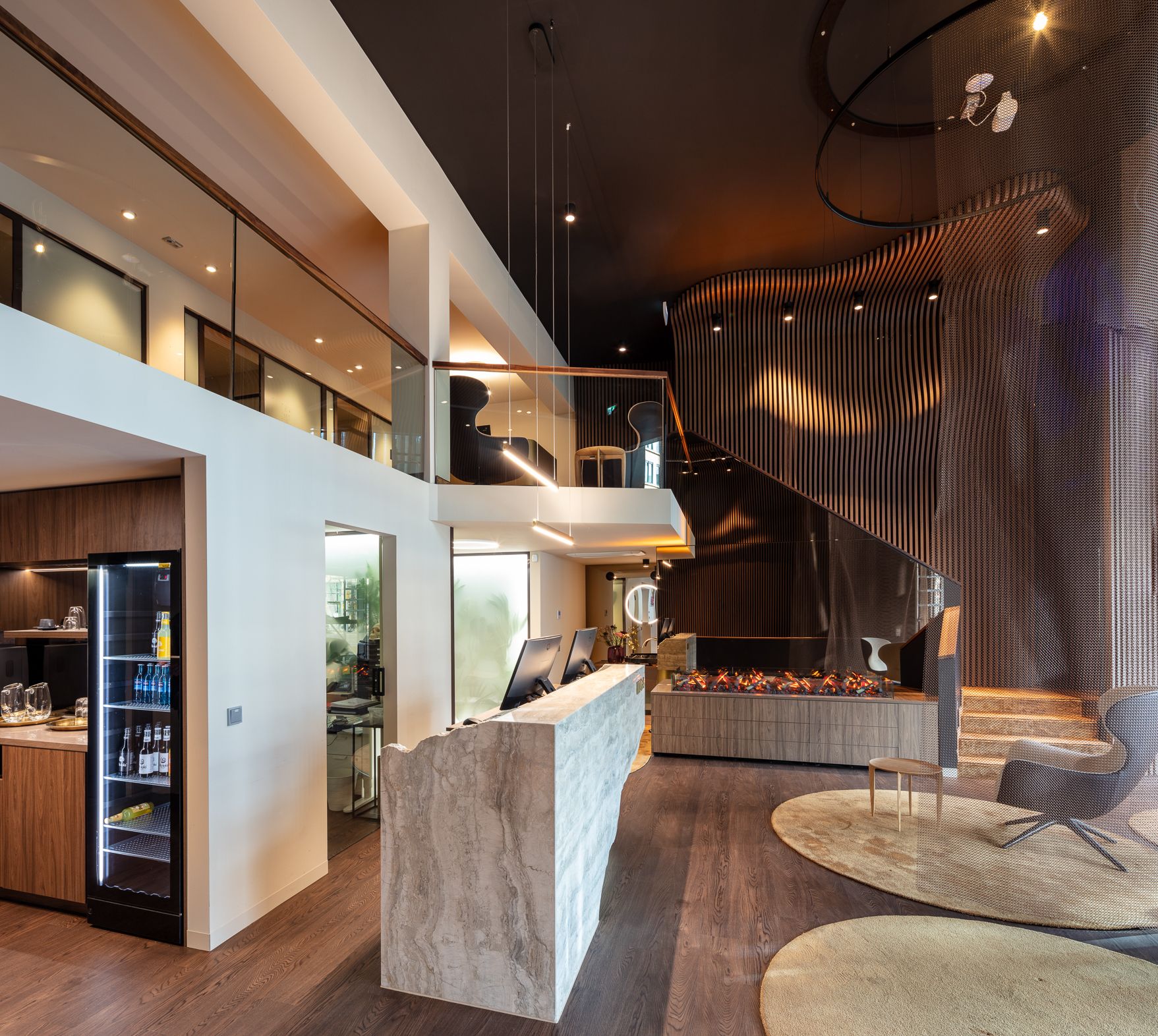 Moderne Lounge mit geschwungenen Holzpaneelen, elektrischem Kamin und runden Sitzbereichen mit grauen Sesseln und halbtransparenten Vorhängen