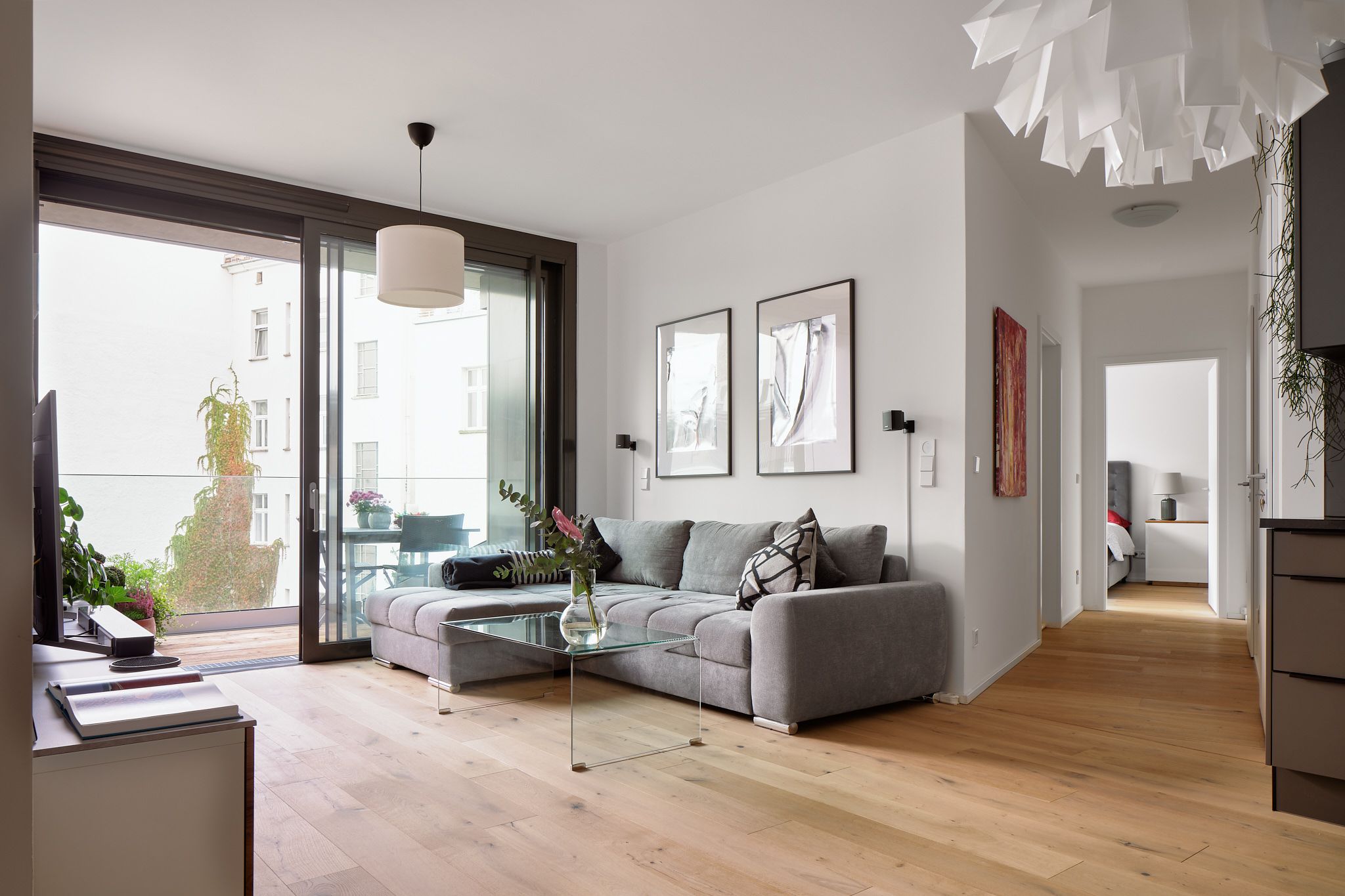 Helles Wohnzimmer mit grauem Ecksofa, Glastisch, zwei Wandbildern und großem Fenster mit Blick auf Balkon und Nachbargebäude
