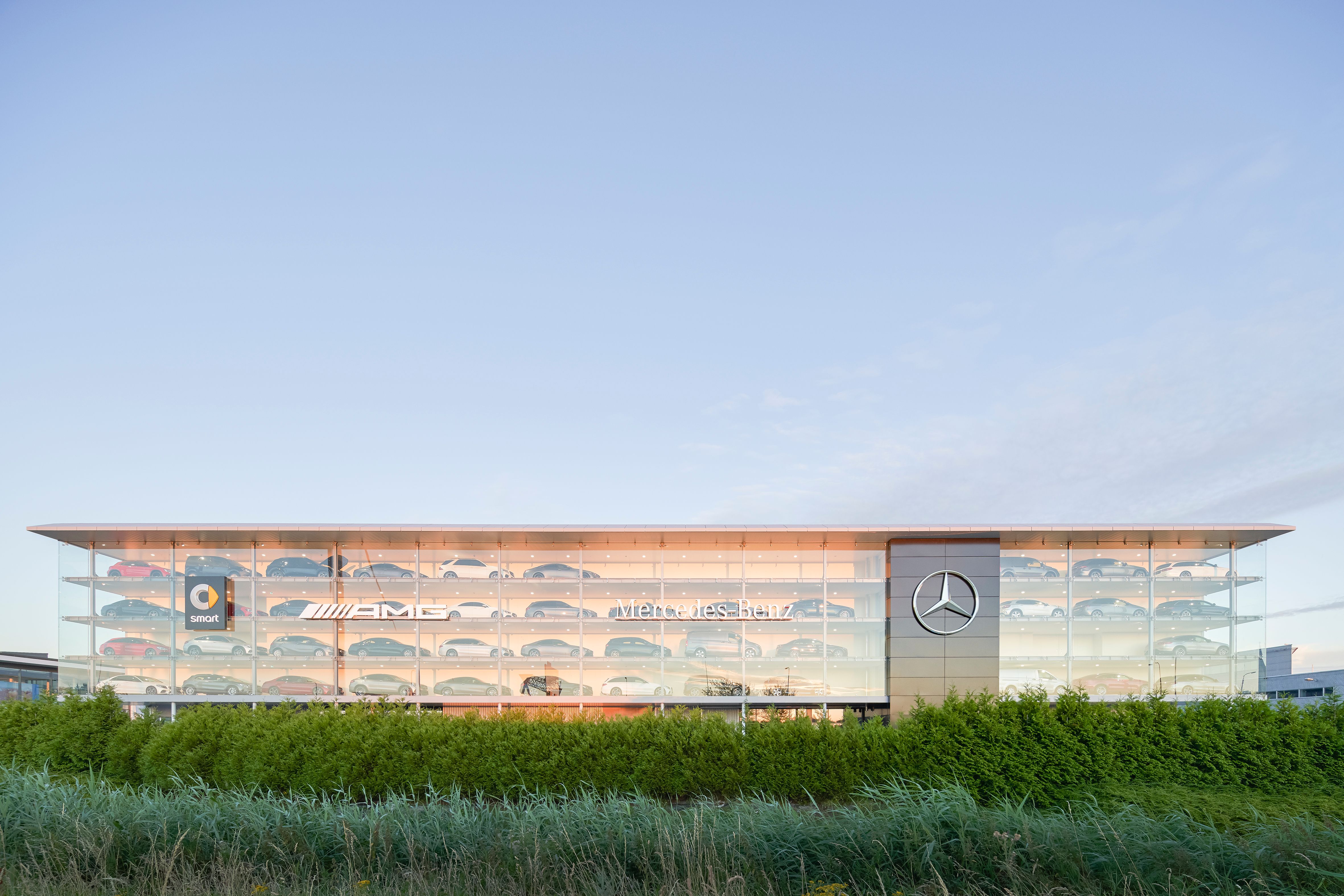 Moderne Glasfassade eines Mercedes-Benz Autohauses mit mehreren Autos in den oberen Etagen, umgeben von Grün.