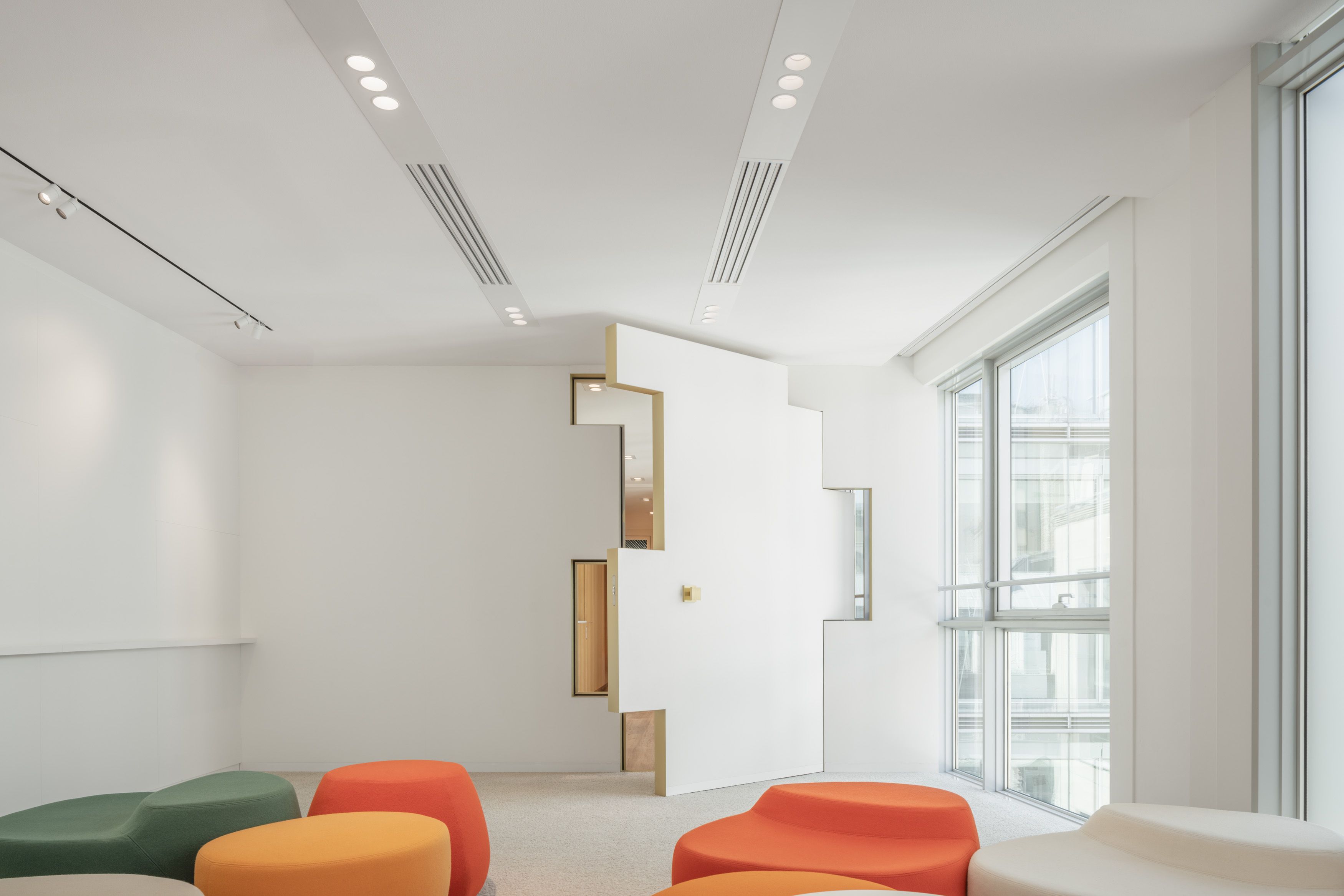 Modernes Interieur mit einer großen, teilweise geöffneten weißen geometrischen Tür, bunten, abgerundeten Ottomane in Orange, Grün und Beige und großen Fenstern auf der rechten Seite.