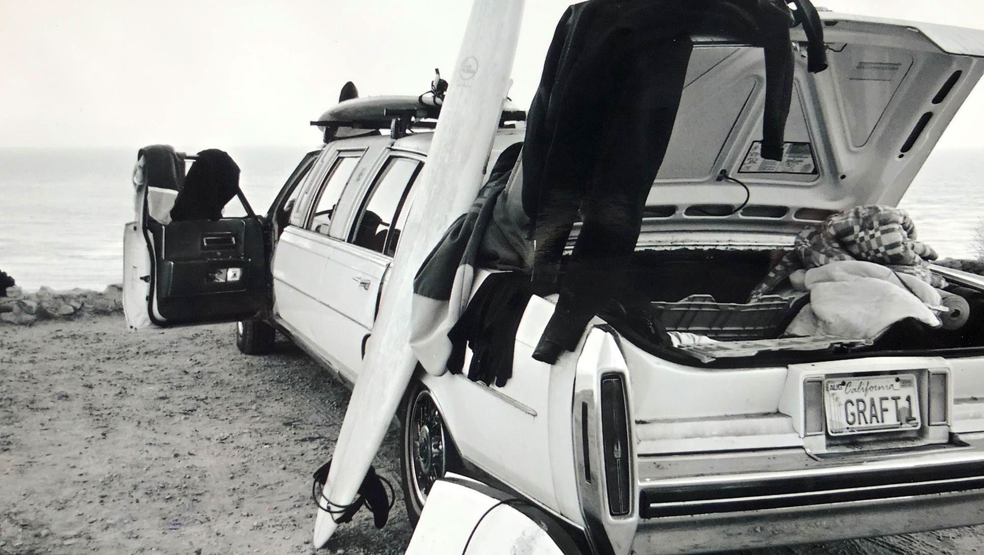 Amerikanisches Auto am Strand mit offenem Kofferraum und offenen Türen. Ein Surfbrett ist ans Auto gelehnt