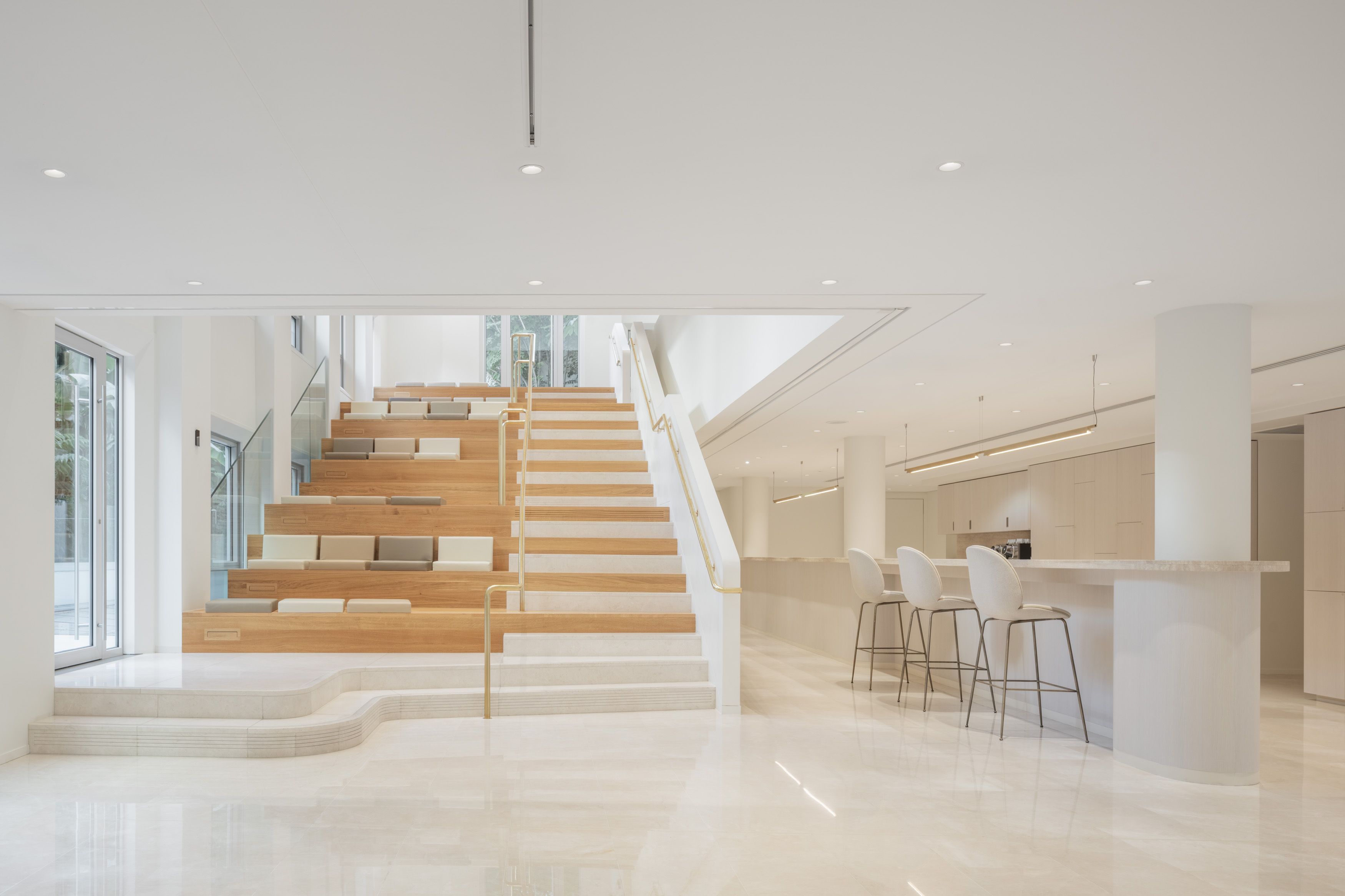 Moderne Innenarchitektur mit einer Treppe aus Holz und weißen Wänden. Links eine Treppe mit Sitzgelegenheiten, rechts eine Küchenzeile mit Barhockern.