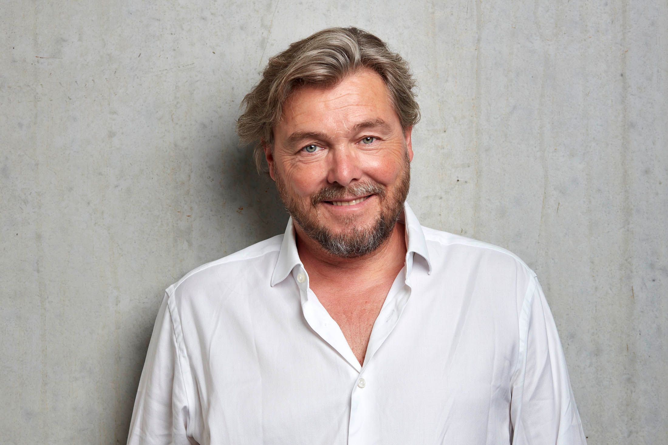 Farbfoto von Lars Krückeberg, er trägt ein weißes Hemd mit offenem Kragen und lächelt entspannt
