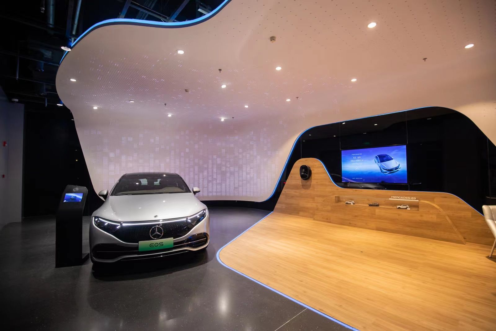 Ein weißes Elektroauto der Marke Mercedes-Benz steht in einem modernen Ausstellungsraum neben einer geschwungenen Holzstruktur mit integriertem Bildschirm.
