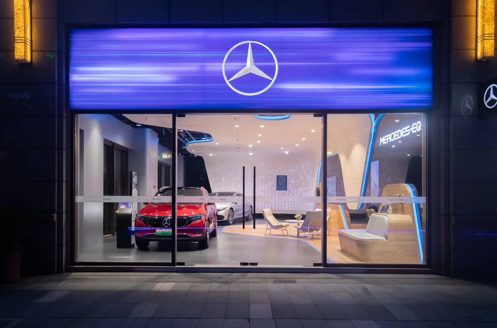 Schaufenster eines Mercedes-Benz-Autohauses mit zwei ausgestellten Fahrzeugen und beleuchtetem Mercedes-Stern-Logo über dem Eingang.