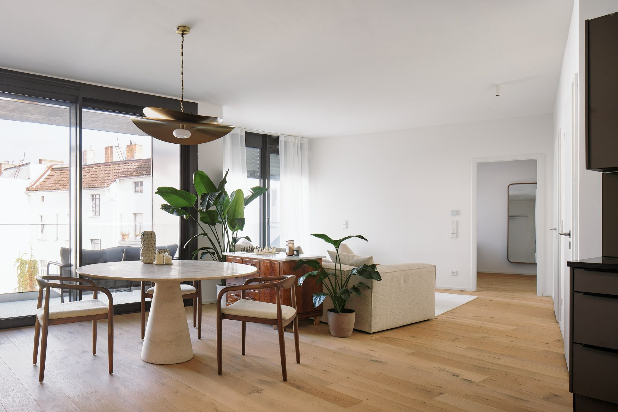 Moderner Wohn- und Essbereich mit großem Fenster, rundem weißen Tisch, Holzstühlen, Pflanzen und hellem Holzboden