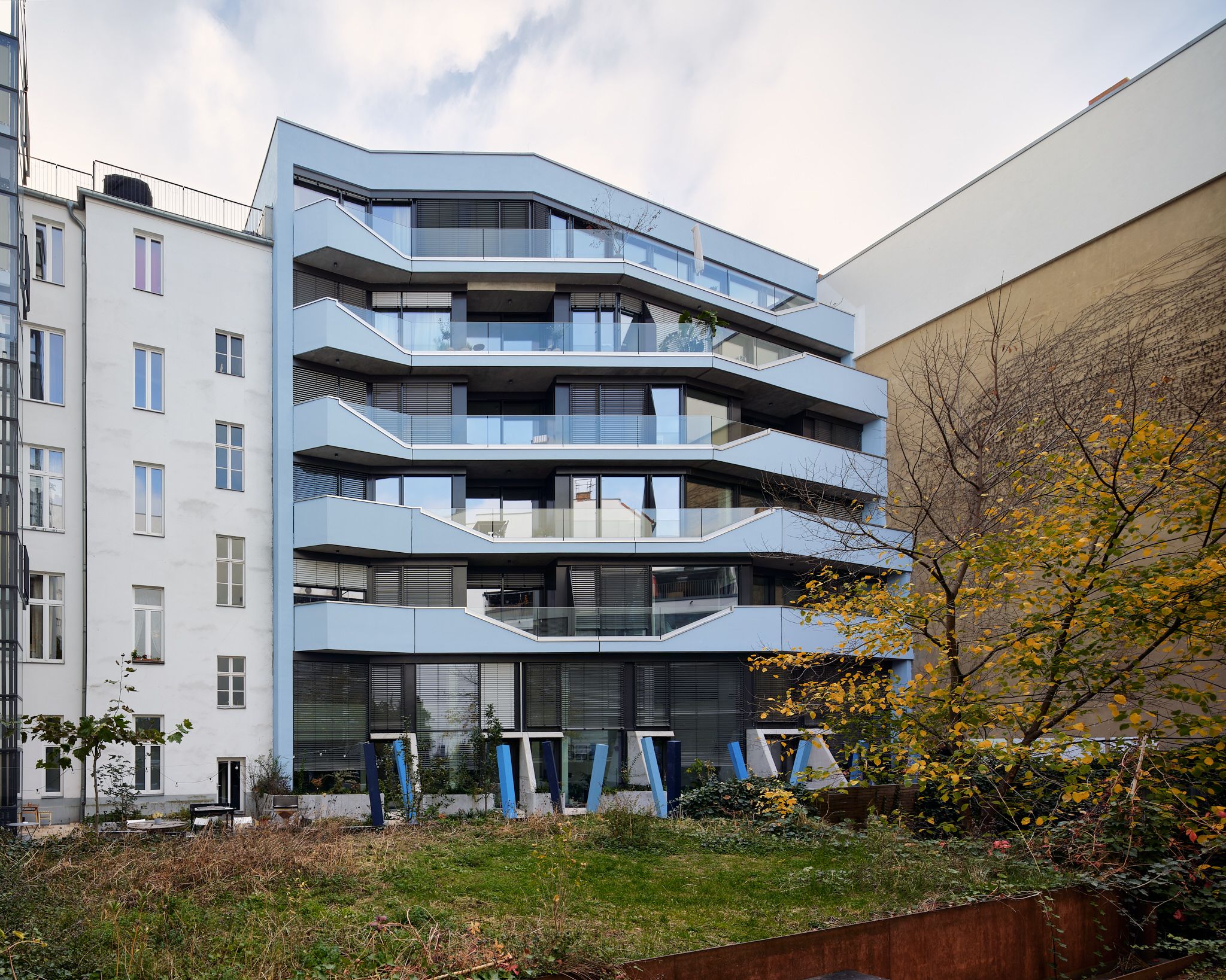 Modern white residential building with staggered balconies and large glass windows, adjacent to older buildings, overlooking a small courtyard with plants and playground equipment