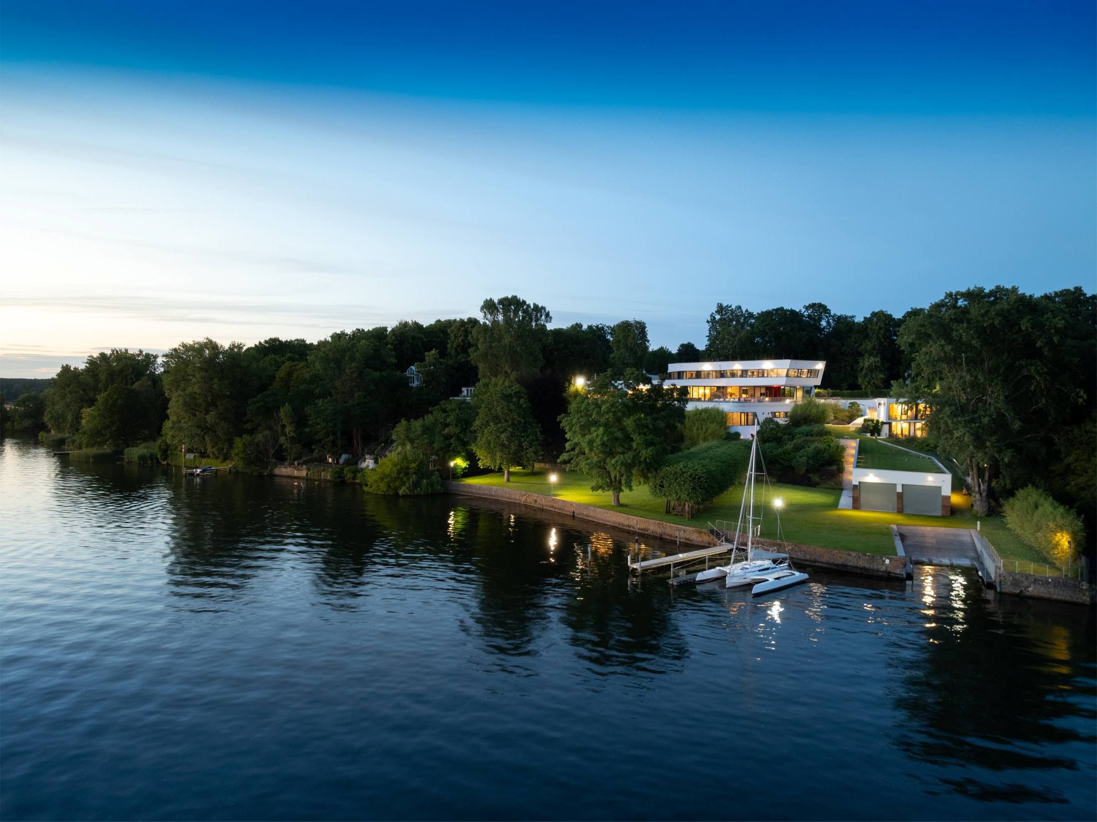 Modern villa with large windows and illuminated garden on the shore of a lake with dock