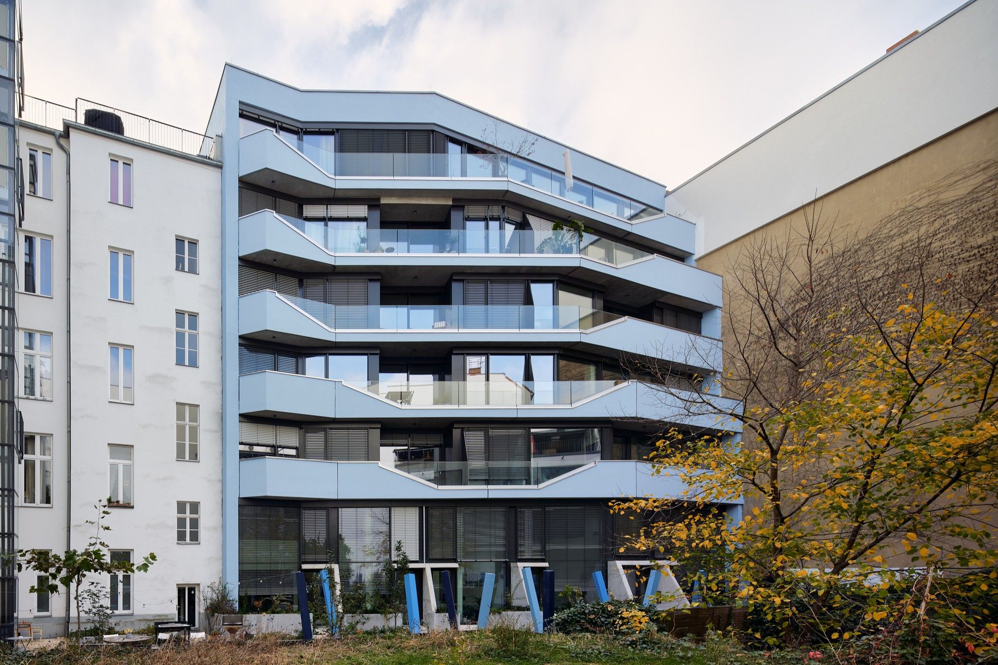 Modern multi-story residential building with angular balconies and large windows next to older buildings and a garden area with small trees and shrubs