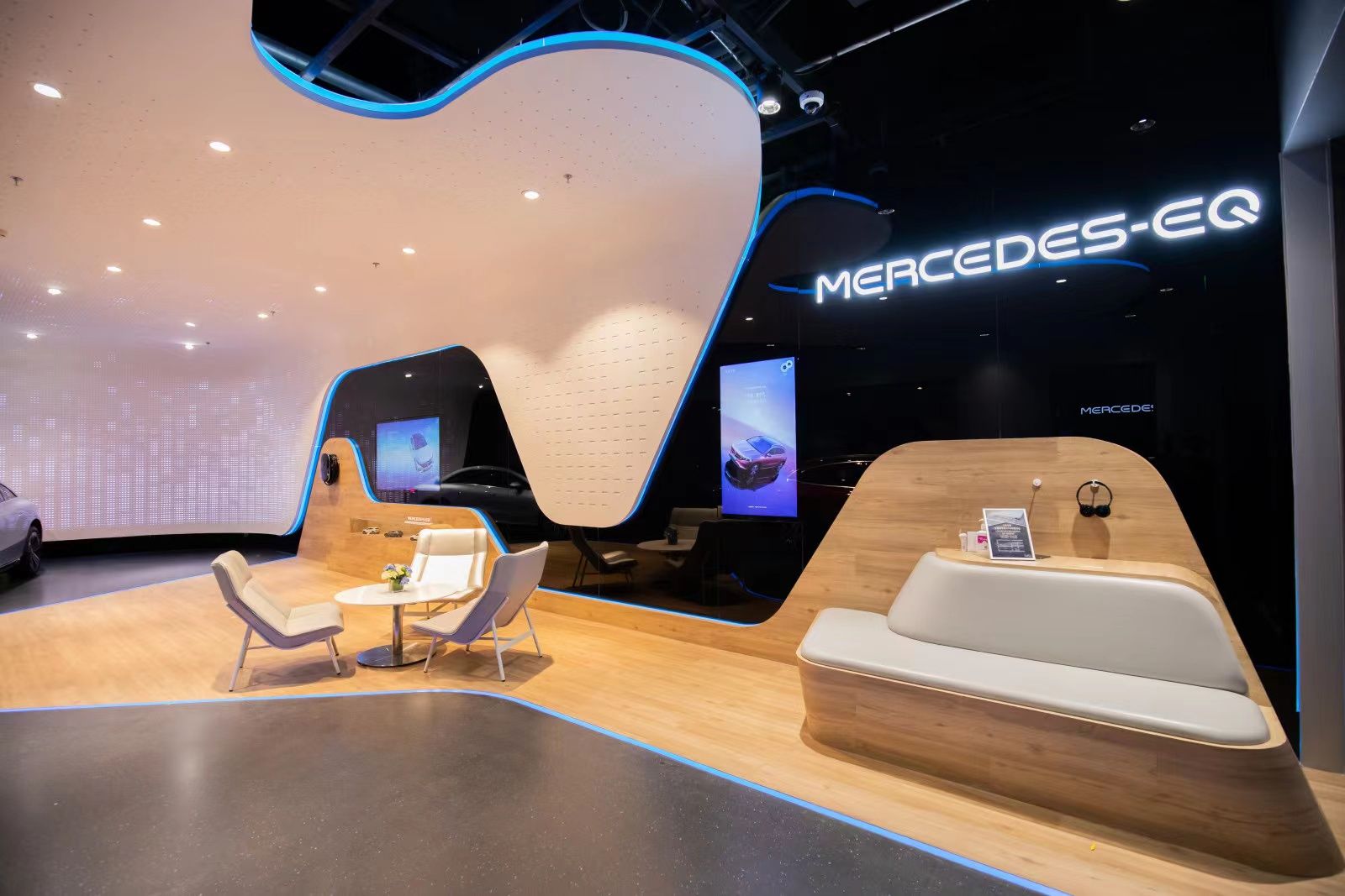 Modern exhibition area with curved wooden and white seating and illuminated 'MERCEDES-EQ' sign on black wall