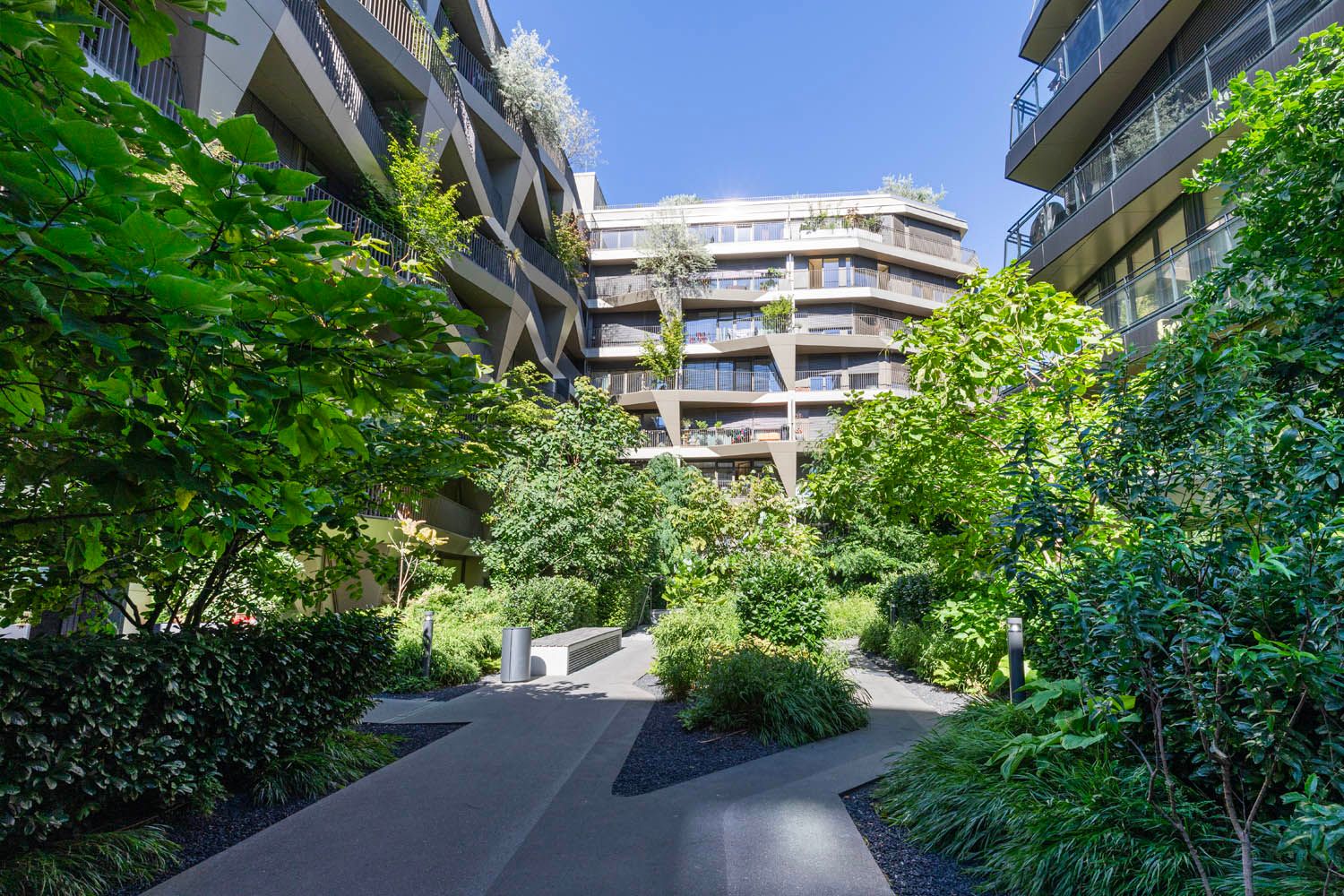 Modern residential buildings surrounding a landscaped courtyard with various green plants and trees under a clear blue sky