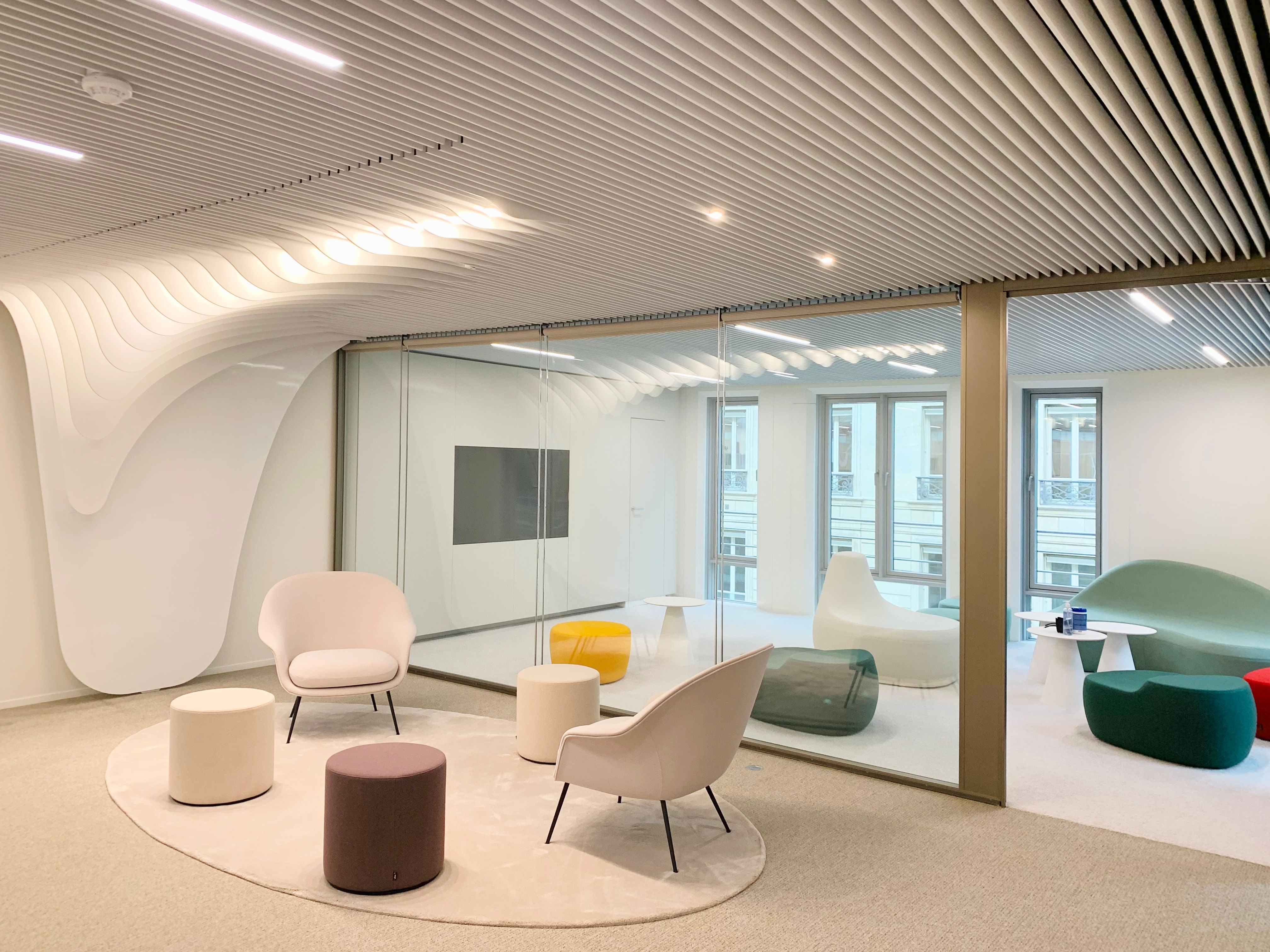 Modern office lounge with a beige armchair and small white side table on a round rug, next to a glass wall enclosing a room with colorful ottomans