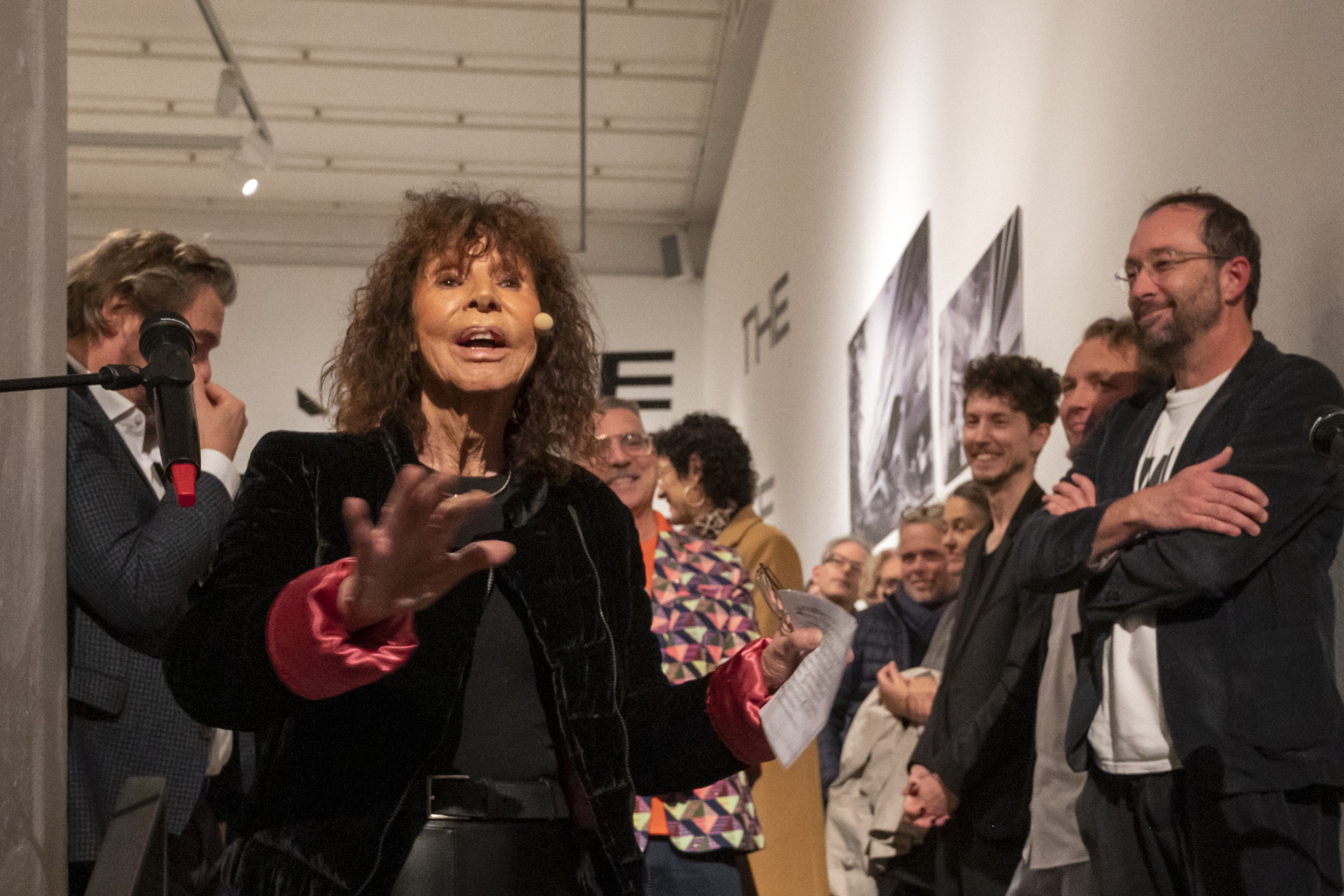 a woman speaks into a mikrophone, several people surround her and listen attentively