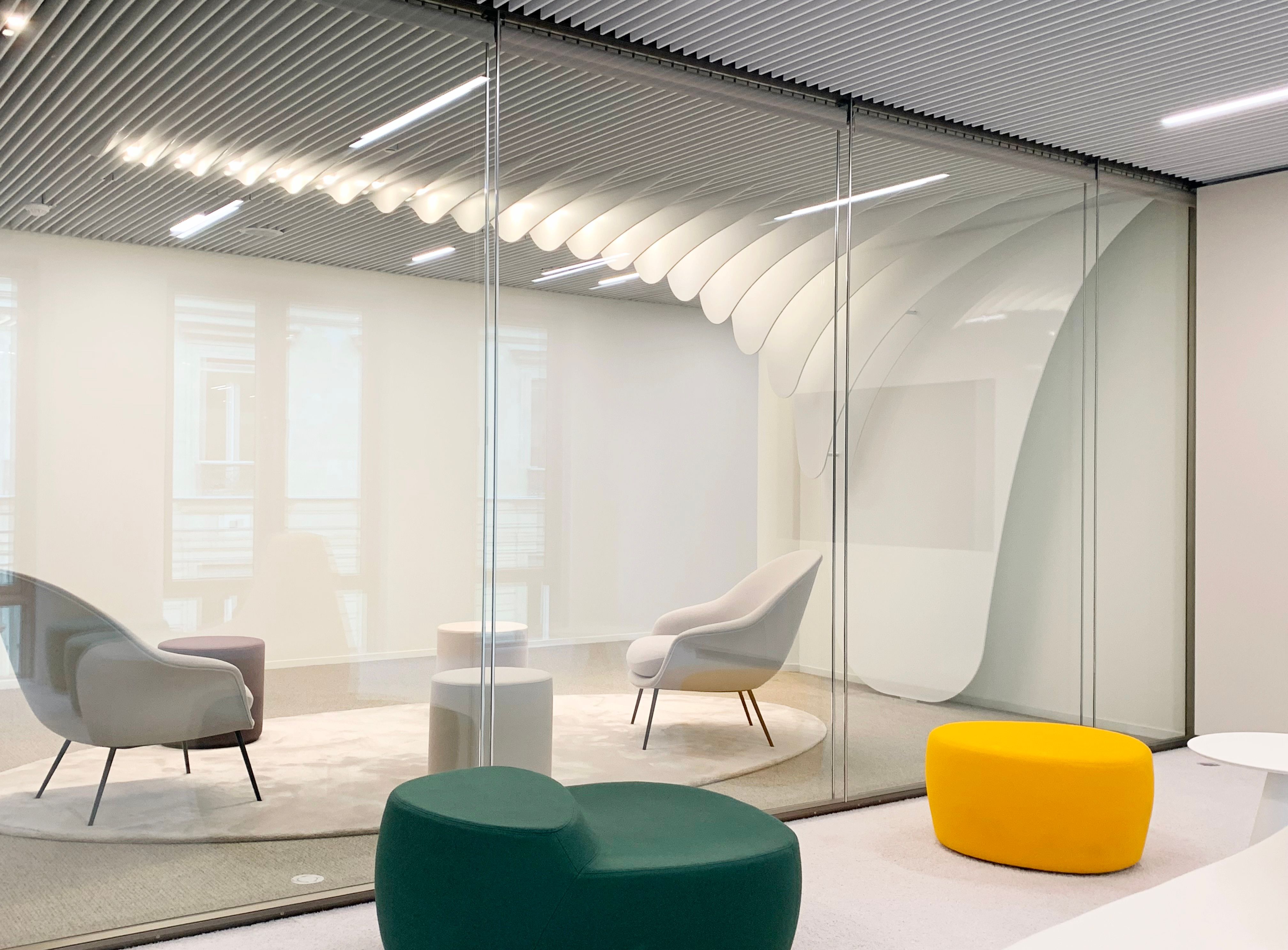 Modern office lounge with a beige armchair and small white side table on a round rug, next to a glass wall enclosing a room with colorful ottomans