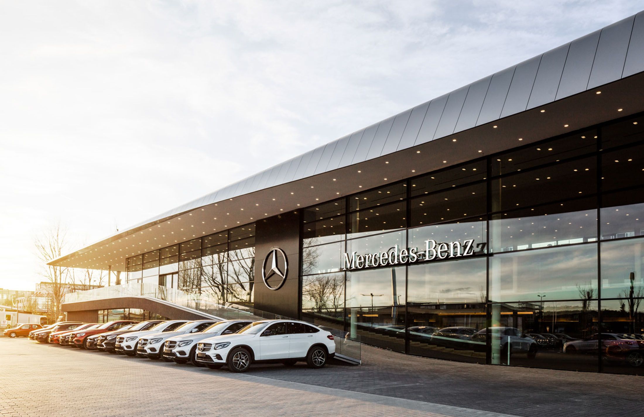 Mercedes-Benz dealership building with large glass windows and logo, row of parked cars outside