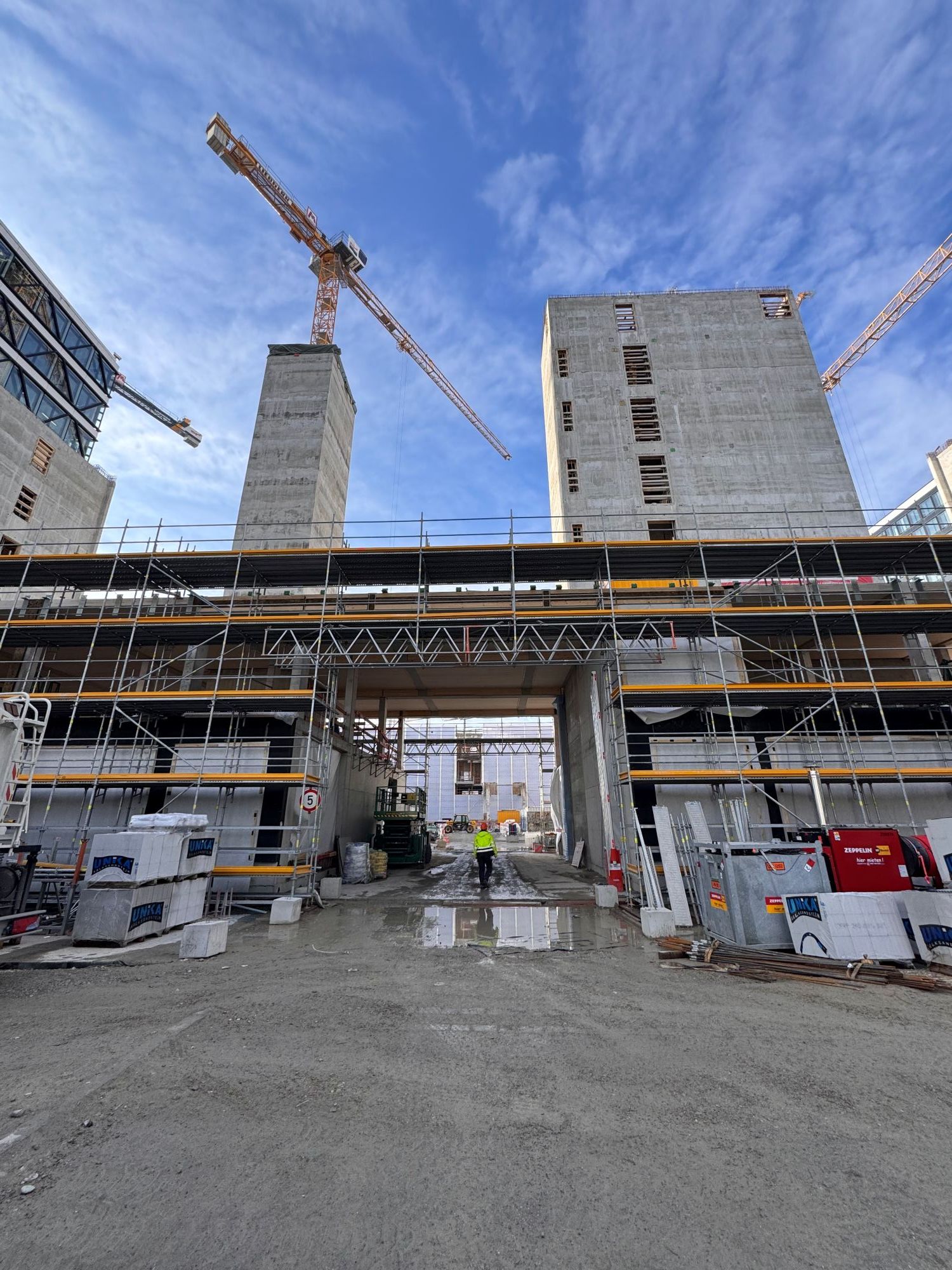 Construction site with several tall concrete buildings, cranes, and a construction worker in a safety vest on unpaved ground