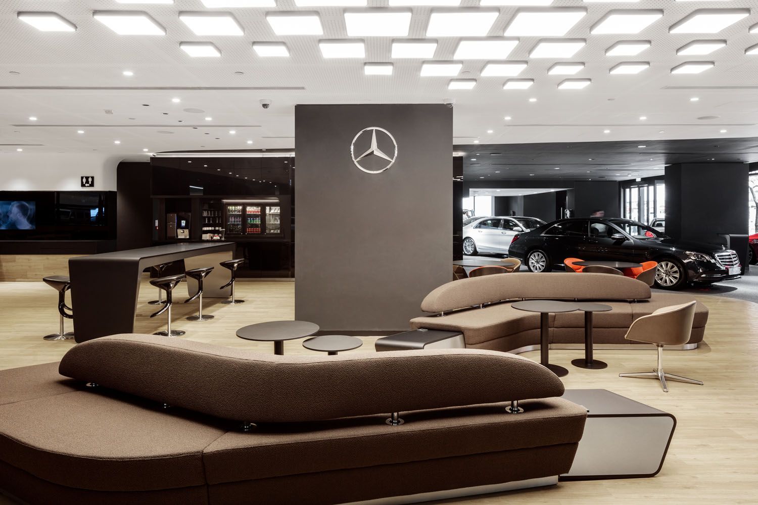 Modern lounge with brown sofas and tables in front of a wall featuring the Mercedes star in a car showroom with several parked cars in the background