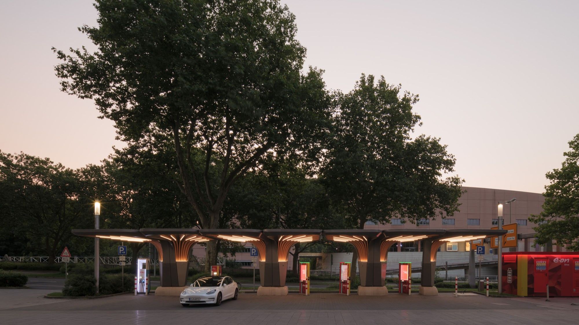 Electric vehicle charging station with multiple chargers under a canopy shaped like tree trunks and a white car parked at one charger