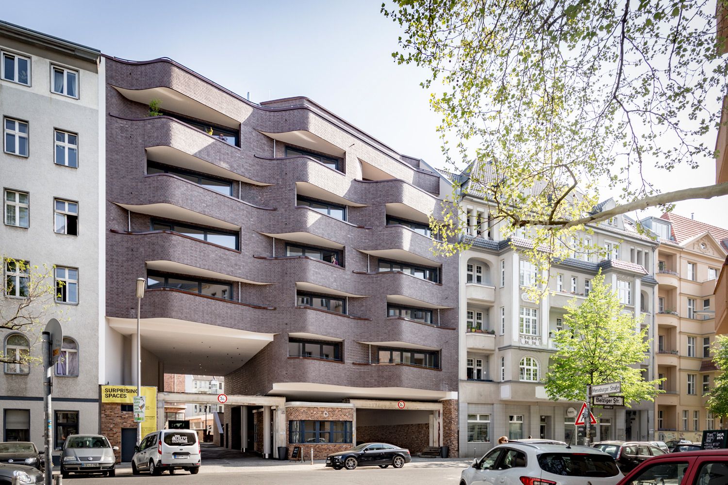 Street view of a modern multi-story building with curved balconies next to older residential buildings and parked cars