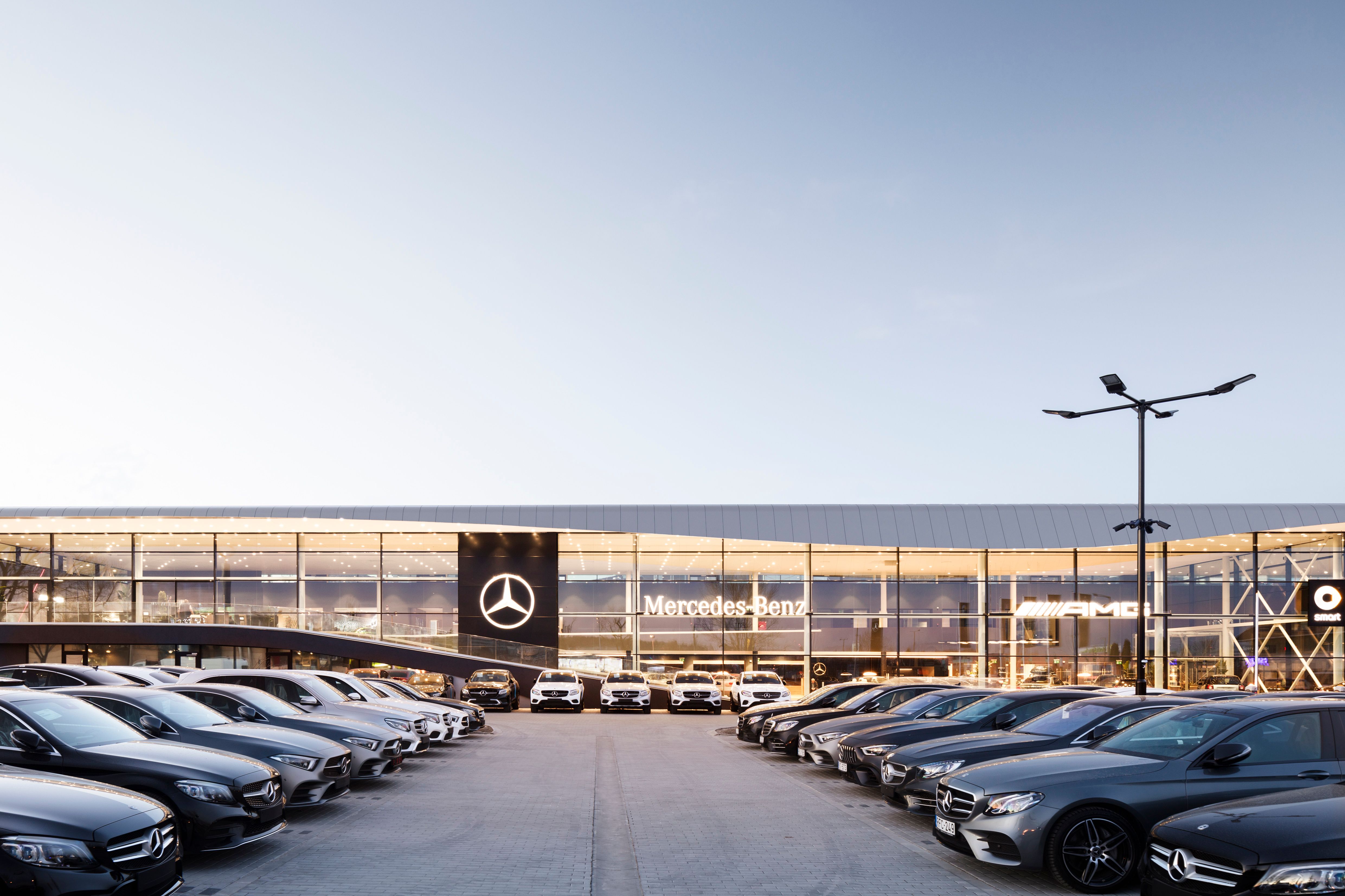 Mercedes-Benz dealership with multiple cars in the parking lot, modern building with glass facade and illuminated logo.