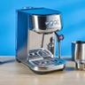 A máquina de espresso Breville Bambino Plus em aço inoxidável prata com acessórios, incluindo jarra de leite, compactador e porta-filtro, exibida extraindo espresso sobre um fundo azul