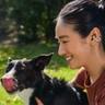 Una giovane donna con i capelli scuri raccolti in una coda di cavallo, che indossa una maglia rosso scuro e un auricolare Shokz OpenDots ONE, coccola il suo cane in un giardino soleggiato mentre il cane si lecca il naso
