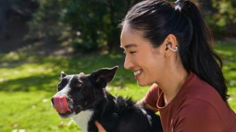 En ung kvinde med mørkt hår sat i hestehale iført en mørkerød top og Shokz OpenDots ONE ørepropper, der hygger sig med sin hund i en solrig have, mens hunden slikker sig om næsen