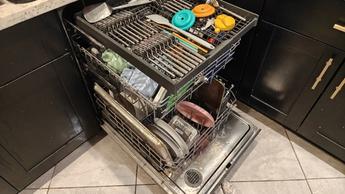 Mesin pencuci piring Whirlpool WDT750SAKZ terbuka, menampilkan piring-piring bersih di dalamnya, dengan lemari dapur hitam dan lantai keramik yang terlihat.