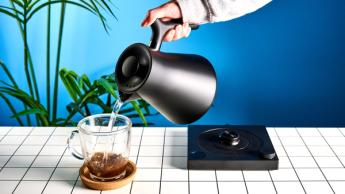 La tetera eléctrica Fellow Corvo EKG en negro mate sobre un fondo azul, acompañada de una taza de vidrio y una mesa en blanco y negro