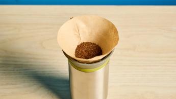 Cafeteira portátil pour-over Wacaco Cuppamoka de aço inoxidável com filtros de papel marrons e tampa de bico em fundo azul